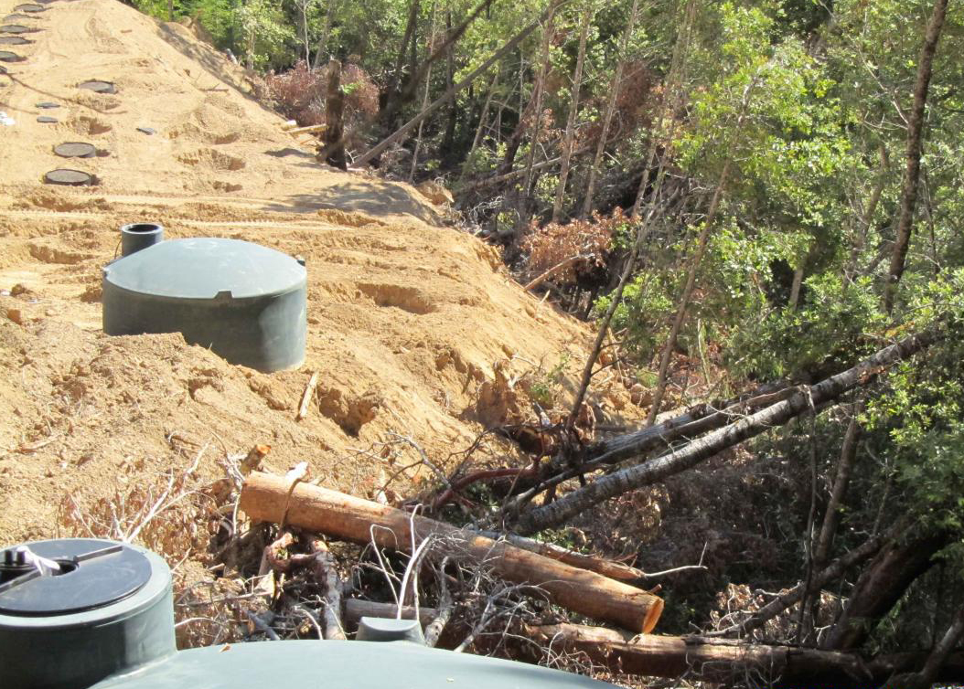 Photograph of a hillside cannabis grow site. The remains felled conifer trees lie on the right and the cleared grow site is steep. Two plastic water storage tanks sit on leveled ground.