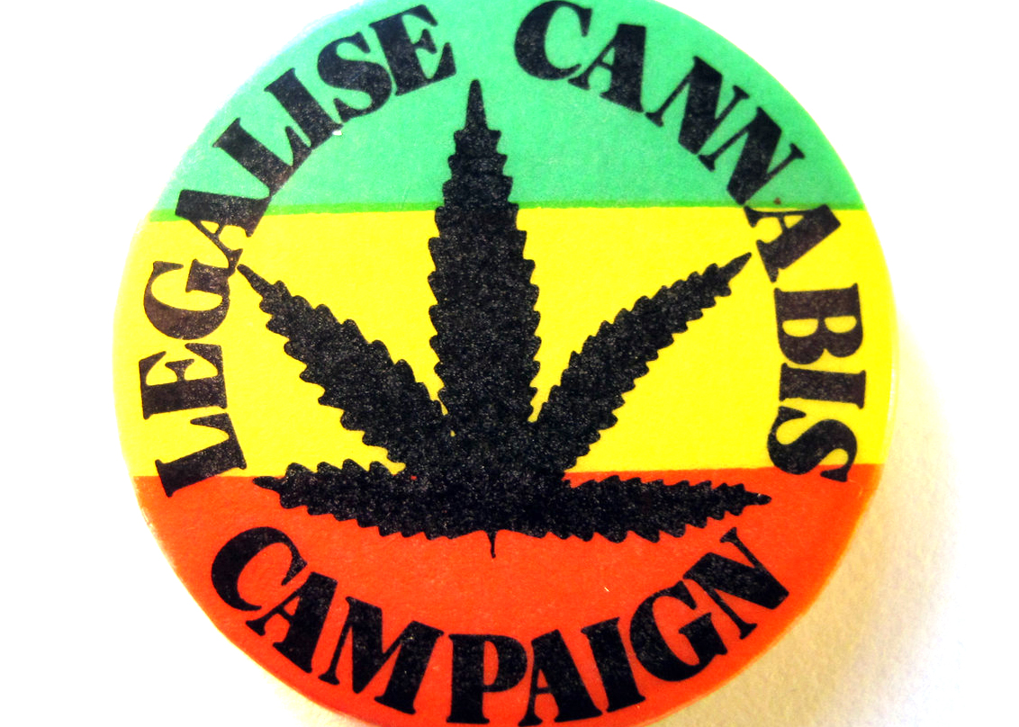 Photo of a button that says "Legalize Cannabis Campaign" arranged in a circle around the silhouette of a cannabis leaf with a striped green, yellow and red background.